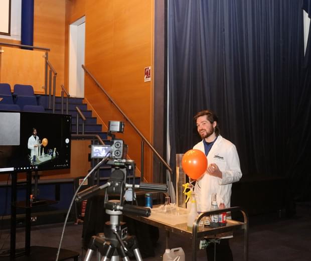 Filming a schools session. A man in a white lab coat is presenting an experiment featuring a big orange baloown. The table in front of him holds various scinetific things like beakers and pots. In the foreground the cameras recording him can be seen