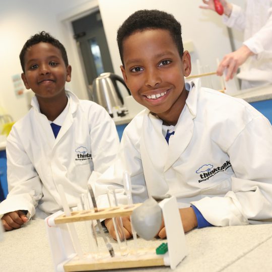 Two children wearing lab coats with test tubes in front of them as part of a schools session.