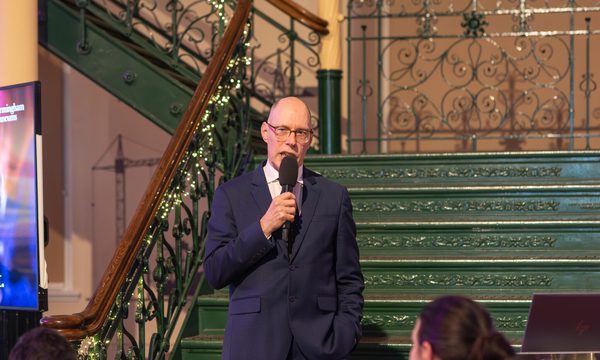 Man in suit speaking into microphone near decorative green staircase with audience nearby.
