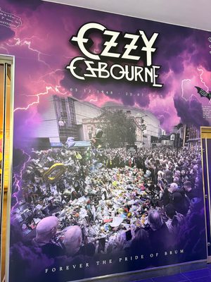 Tribute wall which reads "Ozzy Osbourne 3.12.1948 - 22.7.2025 Forever the pride of Brum. Along side a photo of a crowd of people leaving tributes on near the Black Sabbath bench.