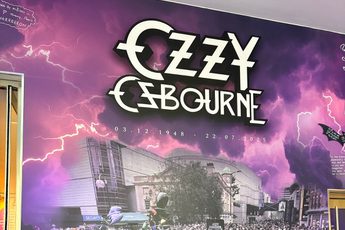 Tribute wall which reads "Ozzy Osbourne 3.12.1948 - 22.7.2025 Forever the pride of Brum. Along side a photo of a crowd of people leaving tributes on near the Black Sabbath bench.