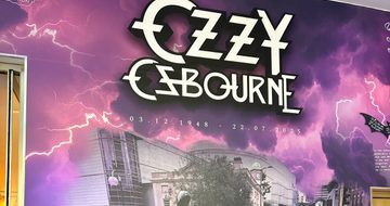 Tribute wall which reads "Ozzy Osbourne 3.12.1948 - 22.7.2025 Forever the pride of Brum. Along side a photo of a crowd of people leaving tributes on near the Black Sabbath bench.