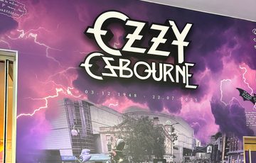 Tribute wall which reads "Ozzy Osbourne 3.12.1948 - 22.7.2025 Forever the pride of Brum. Along side a photo of a crowd of people leaving tributes on near the Black Sabbath bench.