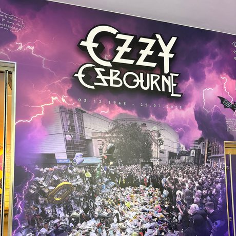 Tribute wall which reads "Ozzy Osbourne 3.12.1948 - 22.7.2025 Forever the pride of Brum. Along side a photo of a crowd of people leaving tributes on near the Black Sabbath bench.