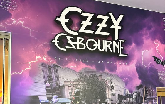 Tribute wall which reads "Ozzy Osbourne 3.12.1948 - 22.7.2025 Forever the pride of Brum. Along side a photo of a crowd of people leaving tributes on near the Black Sabbath bench.