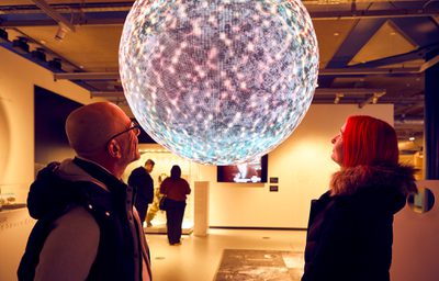 Two people gaze at a large, illuminated, planet in a gallery.