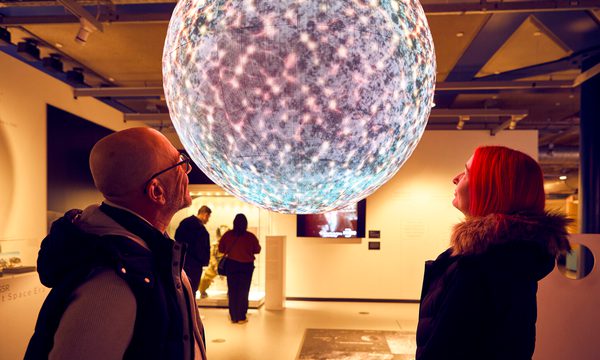 Two people gaze at a large, illuminated, planet in a gallery.