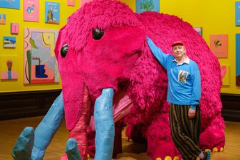 Joe Lycett standing next to a large pink woolly mammoth artwork.