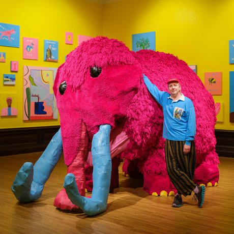 Joe Lycett standing next to a large pink woolly mammoth artwork.