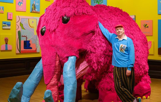 Joe Lycett standing next to a large pink woolly mammoth artwork.