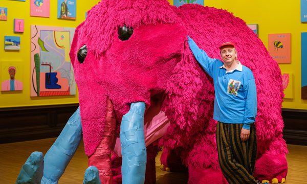 Joe Lycett standing next to a large pink woolly mammoth artwork.