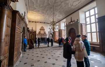 Visitors in the a great hall with stone floor, wood panelled wall and ornate ceiling. A large fireplace is behind the visitors.