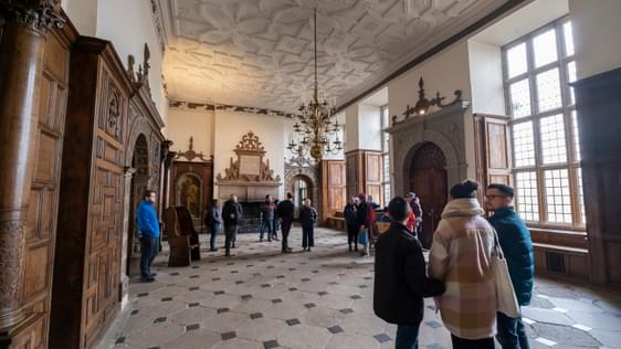 Visitors in the a great hall with stone floor, wood panelled wall and ornate ceiling. A large fireplace is behind the visitors.