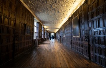 Two visitors in the a Long Gallery with wood panelled walls and ornate ceiling.