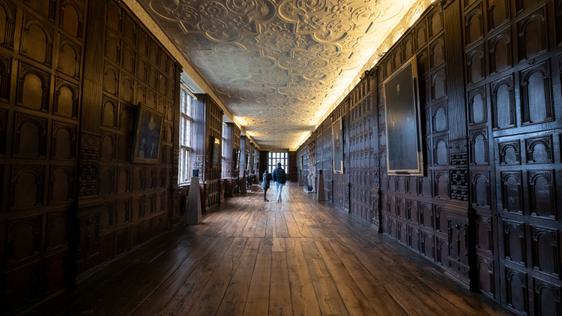 Two visitors in the a Long Gallery with wood panelled walls and ornate ceiling.