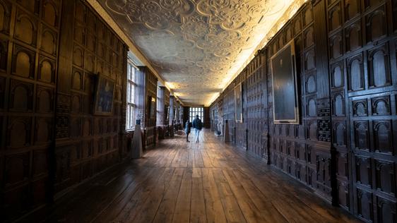 Two visitors in the a Long Gallery with wood panelled walls and ornate ceiling.