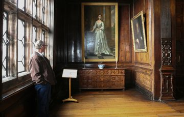 A man leaning against a large leaded window looking at a painting of lady that is hanging on a wood-panlled wall.