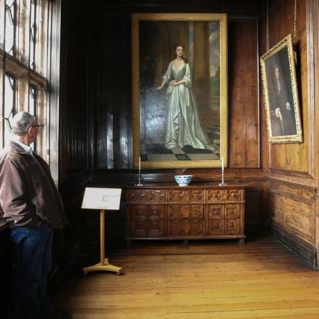 A man leaning against a large leaded window looking at a painting of lady that is hanging on a wood-panlled wall.