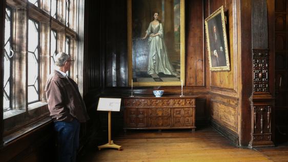 A man leaning against a large leaded window looking at a painting of lady that is hanging on a wood-panlled wall.
