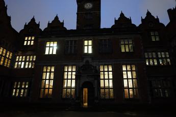 A view of Aston Hall from outside at night. The shape of the hall is a dark silhouette and the windows are lit up by the glowing candlelights from within the rooms.