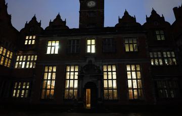A view of Aston Hall from outside at night. The shape of the hall is a dark silhouette and the windows are lit up by the glowing candlelights from within the rooms.