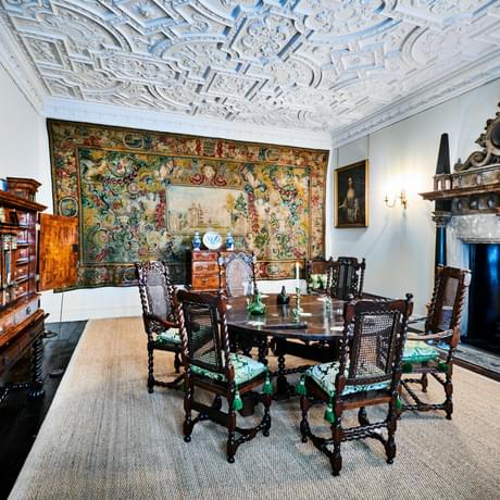 Dining room with wooden table and chairs, ornate plaster ceiling, large fireplace and tapestry on the wall.