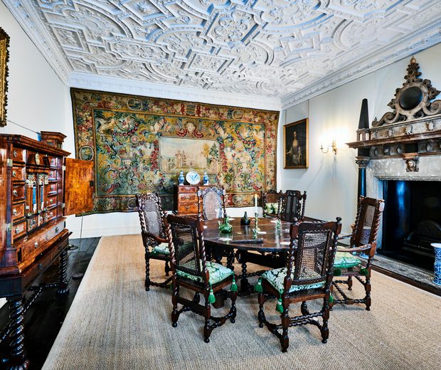 Dining room with wooden table and chairs, ornate plaster ceiling, large fireplace and tapestry on the wall.