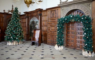 A decorated Christmas tree and garland around a door, in a grand hall with wood panelled walls and paintings on the wall.