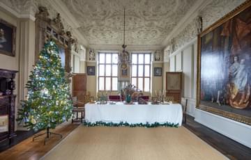 A large grand dining room with ornate ciling, paintings on wall and decorated for Christmas with a tree near a large fireplace.
