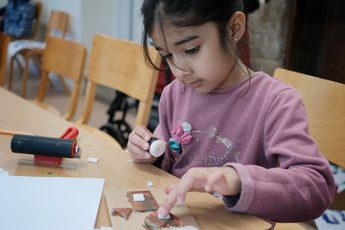 A child is sitting at a table glueing paper on to her artwork.