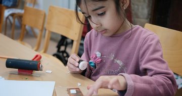 A child is sitting at a table glueing paper on to her artwork.