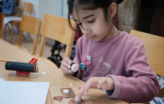 A child is sitting at a table glueing paper on to her artwork.