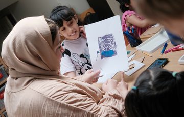 A adult looking at a print that the child has made. The child is smiling in the background.