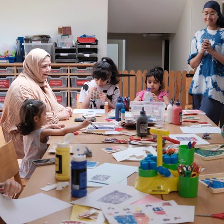 A group of adults and children at a large table which has lots of art supplies on it. The adults are smiling and the children are creating artworks.