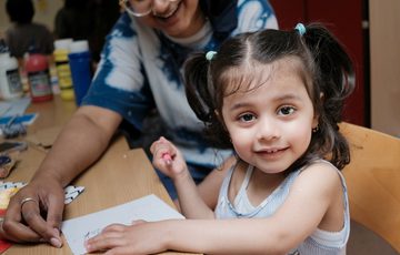A smiling child looks at the camera. She sits at a table and in front of her is a piece of paper. An adult is smiling in the background.
