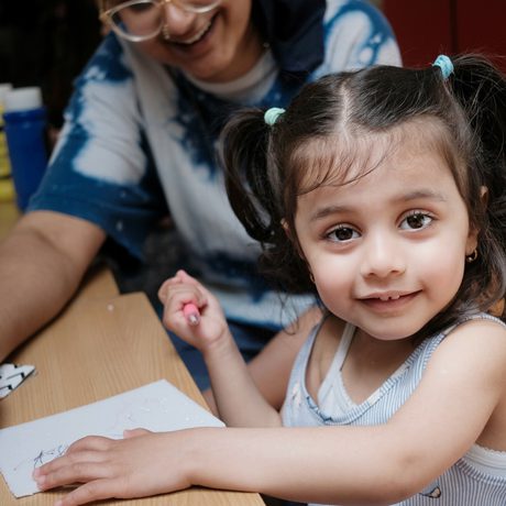 A smiling child looks at the camera. She sits at a table and in front of her is a piece of paper. An adult is smiling in the background.