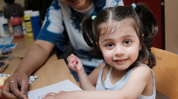 A smiling child looks at the camera. She sits at a table and in front of her is a piece of paper. An adult is smiling in the background.