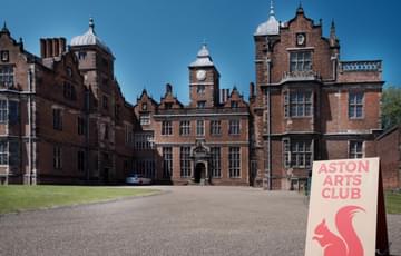 A large red brick Jacobean building and drive. On the drive there is a sign which reads 'Aston Arts Club' and has an illustration of a red squirrel.