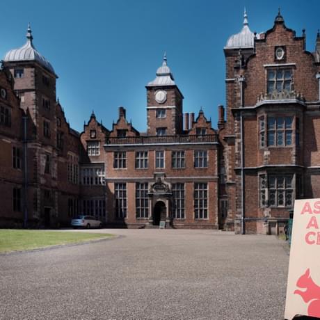 A large red brick Jacobean building and drive. On the drive there is a sign which reads 'Aston Arts Club' and has an illustration of a red squirrel.