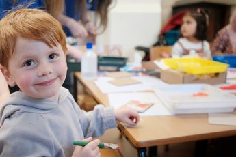 A child is smiling at the camera. He sits at a table creating an artwork, the table has lots of art supplies on it and another child is in the background.