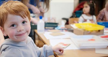 A child is smiling at the camera. He sits at a table creating an artwork, the table has lots of art supplies on it and another child is in the background.
