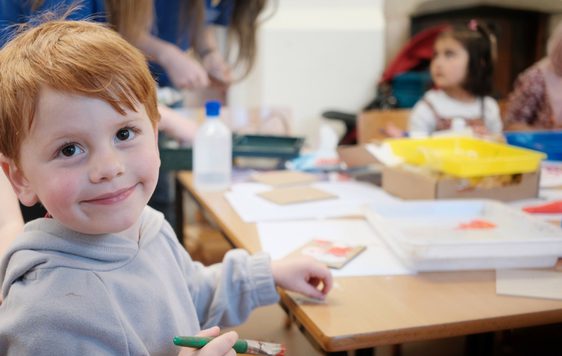 A child is smiling at the camera. He sits at a table creating an artwork, the table has lots of art supplies on it and another child is in the background.