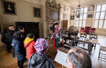 A tour guide dressed as Sir Thomas Holte giving a tour to a group of adults and children. They are in a in Great Dining Room.