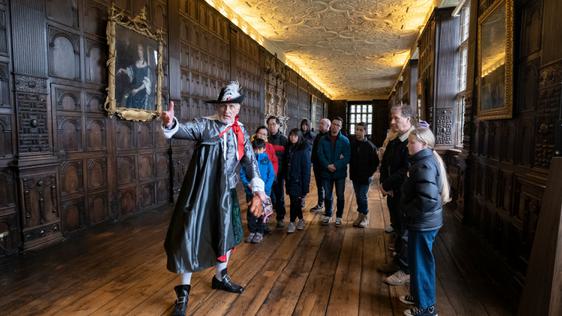 A tour guide dressed as Sir Thomas Holte giving a tour to a group of adults and children. They are in a grand wood panelled Long Gallery.