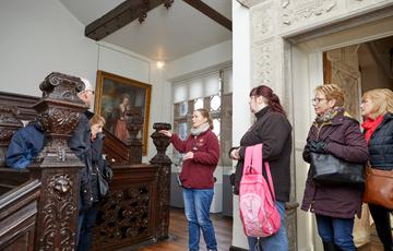A group of adults on a tour of an historic house. They are standing near an intricate wooden staircase and door.