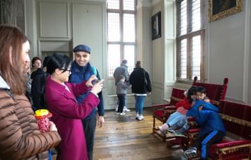 A family taking a photo of children sitting on bench inside a Jacobean room with wooden floors and large tall windows.