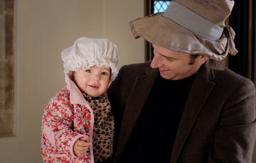 An adult holding a toddler, they are both smiling and wearing historical hats.