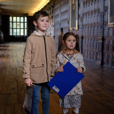 Two children stand in the long gallery which is a long wood panelled room. The children are holding clipboards and pencils.