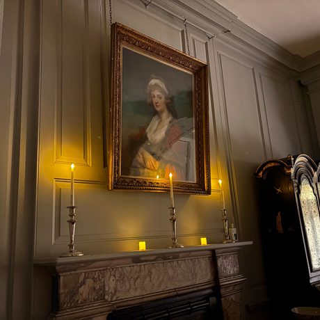 Dimly lit vintage room with portrait, candles on mantel, and ornate wooden cabinet.