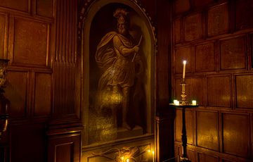 Dimly lit room with wooden paneling, a large knight painting, and candles.